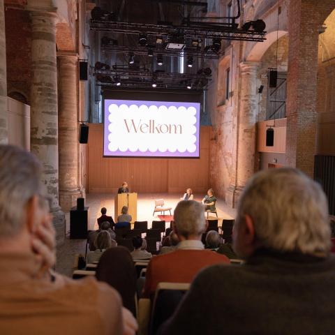 Vrijwilligers kijken toe vanuit de tribune naar het podium van de Predikherenkerk tijdens welkomstwoord van schepen Geypen op de nieuwjaarsreceptie.