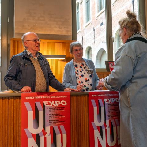 Twee vrijwilligers staan achter een balie met daarop posters van de U, nu!-expo. Ze heten een dame welkom die voor de balie staat.