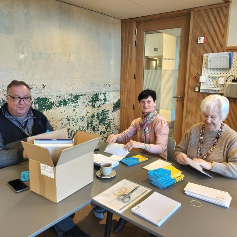 Drie vrijwilligers steken folders van een Soirée MuzE-lezingenreeks in enveloppen en maken deze klaar voor verdeling.