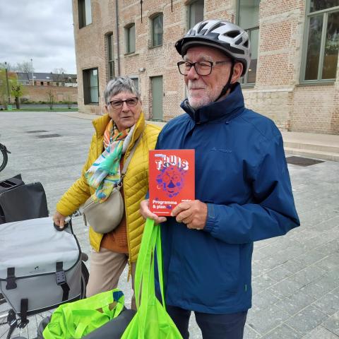 Twee vrijwilligers poseren bij hun fiets. In de fietstassen zitten zakken vol folders. Een vrijwilliger draagt een helm en poseert met een folder van Erfgoeddag.