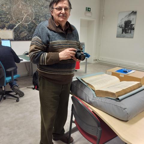 Vrijwilliger poseert met camera in de hand voor een tafel met daarop een groot, oud en dik boek dat is opengeslagen.