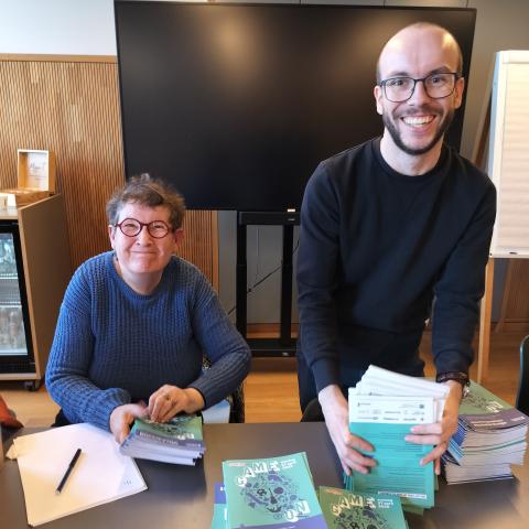Vrijwilliger en medewerker van Erfgoedcel Mechelen poseren voor de camera terwijl ze folders tellen en verdelen in gelijke stapels.